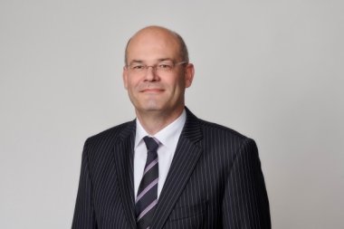 Ein Herr mit sehr kurzem Haar, Brille, einem weißen Hemd, schwarz lilaner Krawatte und einem schwarzen Sacko